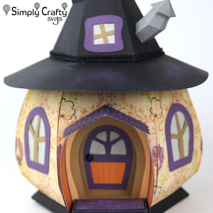 Peut inclure: Une maison de sorci&egrave;re en papier avec un chapeau noir, une porte violette et deux fen&ecirc;tres. La maison est d&eacute;cor&eacute;e d'un motif floral et poss&egrave;de une chemin&eacute;e.