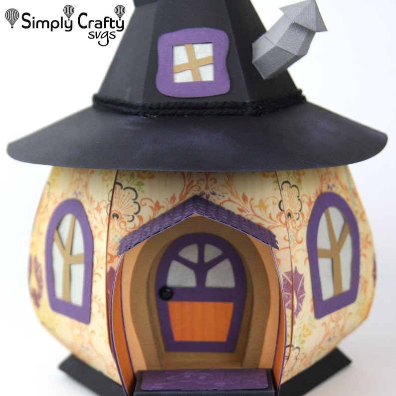 Peut inclure: Une maison de sorci&egrave;re en papier avec un chapeau noir, une porte violette et deux fen&ecirc;tres. La maison est d&eacute;cor&eacute;e d'un motif floral et poss&egrave;de une chemin&eacute;e.