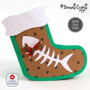 May include: A Christmas stocking-shaped craft project with a fish bone design. The stocking is brown with paw prints, green trim, and a white top. A red and green bow adorns the fish bone. The text "Simply Crafty SVGs" and "SVG Cut File" are visible.