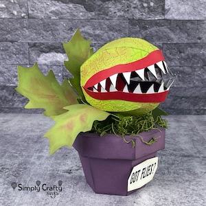 May include: A papercraft model of a carnivorous plant, a Venus flytrap, with a green, textured body, red lips, and white teeth. The plant is in a purple pot with a white label that reads "GOT FLIES?"