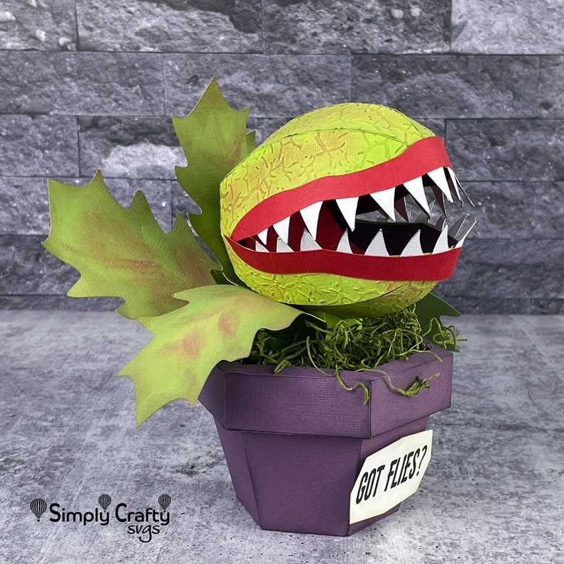 May include: A papercraft model of a carnivorous plant, a Venus flytrap, with a green, textured body, red lips, and white teeth. The plant is in a purple pot with a white label that reads "GOT FLIES?"