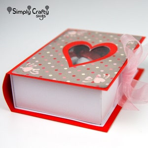 May include: A red and white book-shaped gift box with a heart-shaped window. The cover is gray with pink polka dots and paw prints. A pink ribbon is tied around the box. The text "Simply Crafty svgs" is in the upper left corner.