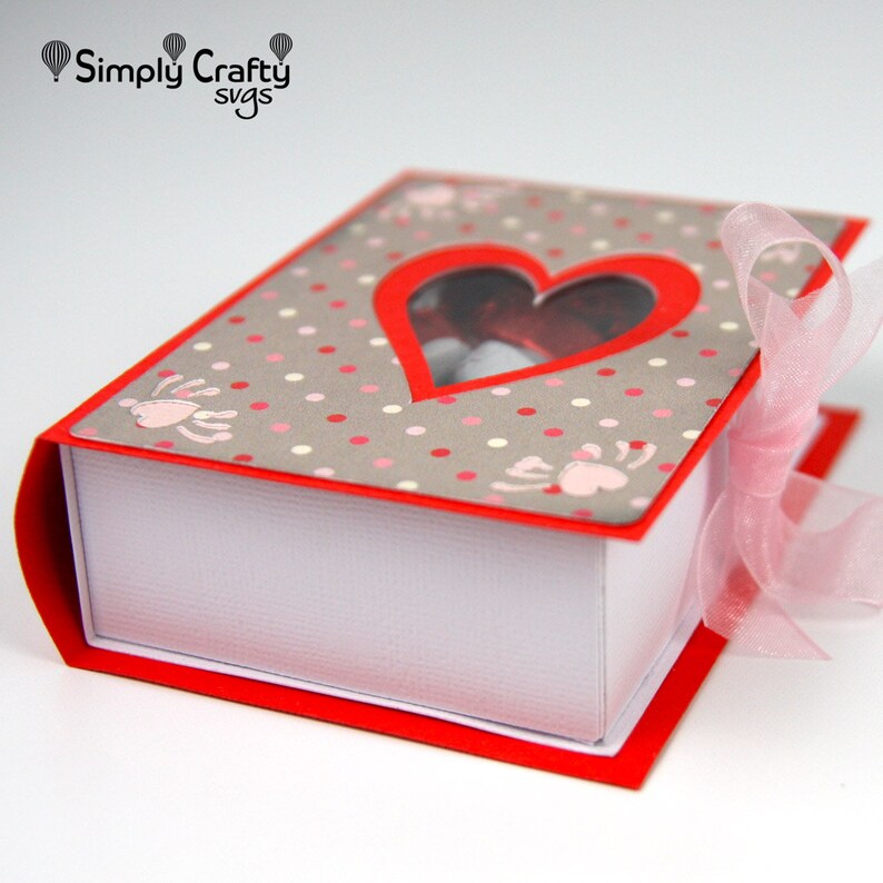 May include: A red and white book-shaped gift box with a heart-shaped window. The cover is gray with pink polka dots and paw prints. A pink ribbon is tied around the box. The text "Simply Crafty svgs" is in the upper left corner.