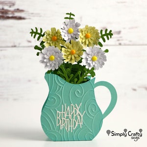 K&ouml;nnte beinhalten: Eine tealblaue Papierhandwerkskrugvase mit einer wei&szlig;en Aufschrift "Happy Birthday" und einem Strau&szlig; wei&szlig;er und gelber Papierblumen.