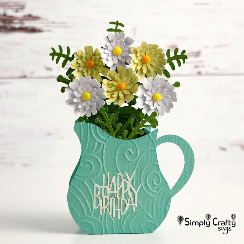 K&ouml;nnte beinhalten: Eine tealblaue Papierhandwerkskrugvase mit einer wei&szlig;en Aufschrift "Happy Birthday" und einem Strau&szlig; wei&szlig;er und gelber Papierblumen.