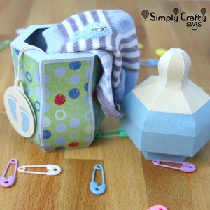 Puede incluir: Un calcet&iacute;n a rayas azul y blanco se encuentra dentro de una caja de regalo con estampado verde y azul con una etiqueta azul con forma de huella. Una caja de regalo con forma de biber&oacute;n azul y blanco se encuentra junto a la caja. Hay varios imperdibles esparcidos sobre una superficie de madera.