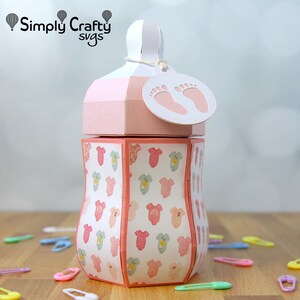 Puede incluir: Caja con forma de biber&oacute;n rosa y blanca con una etiqueta blanca que dice "Simply Crafty svgs" y tiene un dise&ntilde;o de huella. La caja est&aacute; decorada con un patr&oacute;n de bodies de beb&eacute; rosas y verdes.