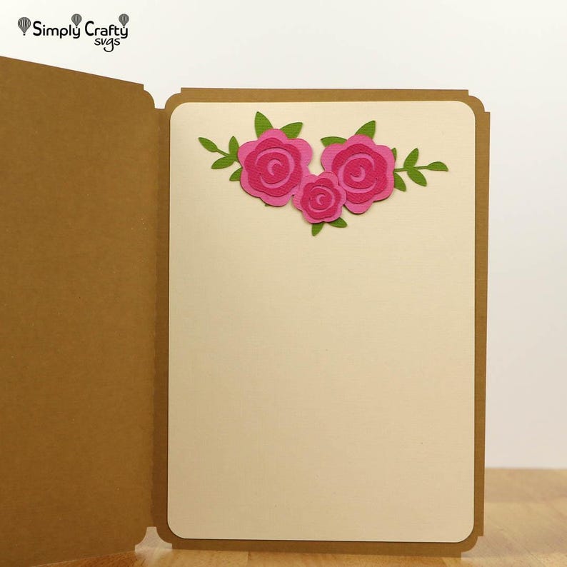 May include: A handmade greeting card with a tan cover and a cream-colored interior. The card features a floral design of three pink roses with green leaves. The text "Simply Crafty svgs" is in the upper left corner.