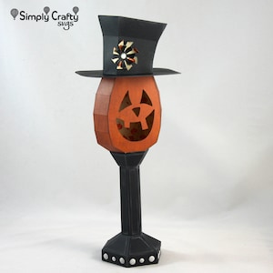 May include: A black and orange papercraft jack-o'-lantern with a black top hat. The jack-o'-lantern has a carved face and is on a black stand with a white polka dot pattern.