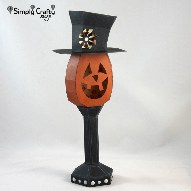 May include: A black and orange papercraft jack-o'-lantern with a black top hat. The jack-o'-lantern has a carved face and is on a black stand with a white polka dot pattern.
