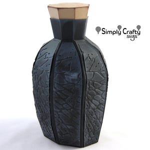 May include: A dark gray, multi-faceted paper craft bottle with a textured, stone-like surface and a light brown hexagonal lid. The text Simply Crafty SVGs is visible in the upper right corner, accompanied by three small hot air balloon icons.