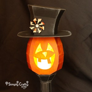 May include: A papercraft jack-o'-lantern with a black top hat and a white and red paper flower. The jack-o'-lantern has a bright orange face with a wide smile and a yellow glow.