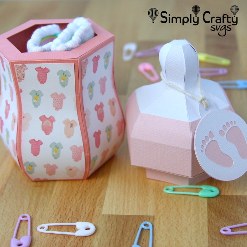 Puede incluir: Dos cajas de regalo para baby shower de color rosa y blanco. Una caja tiene forma de biber&oacute;n y lleva una etiqueta blanca con una huella de pie rosa. La otra caja tiene forma de jarr&oacute;n y tiene un patr&oacute;n rosa y blanco con bodies de beb&eacute;. El texto "Simply Crafty svgs" es visible en la esquina superior derecha de la imagen.