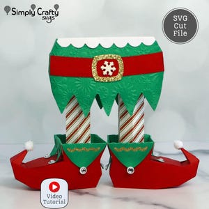 May include: A festive Christmas elf decoration with red shoes, green and red striped legs, and a green top with a red belt. The design includes a gold buckle with a snowflake. The image also includes the text "SVG Cut File" and "Video Tutorial".