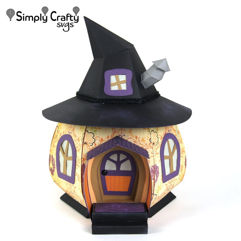 Peut inclure: Une maison de sorci&egrave;re en forme de citrouille avec un chapeau de sorci&egrave;re noir sur le dessus. La maison a une porte violette avec une poign&eacute;e de porte brune, un petit porche avec des marches et deux fen&ecirc;tres avec des cadres violets. La maison est d&eacute;cor&eacute;e d'un motif floral.