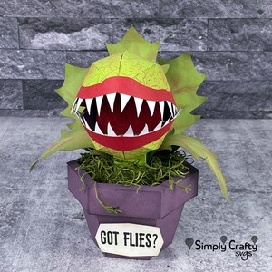 May include: A papercraft model of a carnivorous plant, the Venus Flytrap, in a purple pot. The plant has a large, open mouth with sharp teeth and a red tongue. The pot has a sign that reads "GOT FLIES?" and a small logo for "Simply Crafty SVGs".