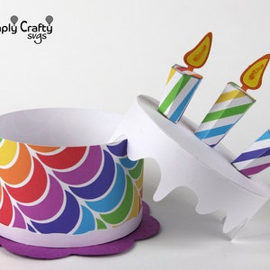 May include: A rainbow-coloured paper cake box with a purple base and three striped candles with orange flames. The box is open and the top is tilted to the side.