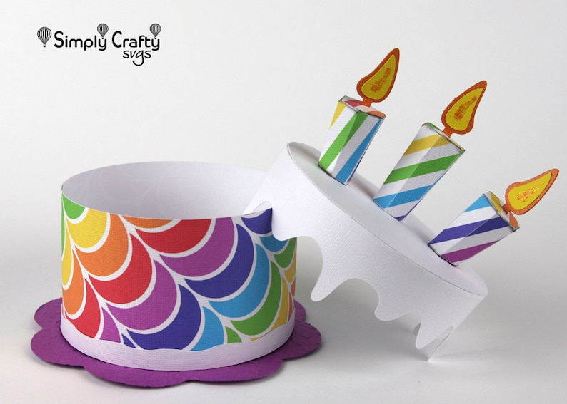 May include: A rainbow-coloured paper cake box with a purple base and three striped candles with orange flames. The box is open and the top is tilted to the side.