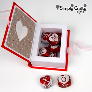 May include: A red and white paper box with a heart-shaped cutout and polka dot pattern, filled with heart-shaped chocolates. The chocolates are wrapped in red foil with white lettering. A pink ribbon is attached.