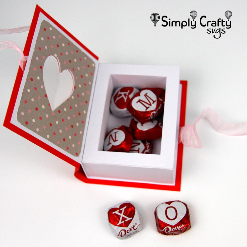 May include: A red and white paper box with a heart-shaped cutout and polka dot pattern, filled with heart-shaped chocolates. The chocolates are wrapped in red foil with white lettering. A pink ribbon is attached.