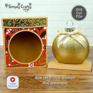 Puede incluir: Un adorno de Navidad dorado con purpurina, con una cinta blanca y una caja de cartón marrón con un diseño estampado en rojo y dorado. La caja está diseñada para contener un adorno de disco de 5 pulgadas (120-126 mm). El texto "Box for 5" (120-126 mm) disc ornament" está impreso debajo de la caja. El texto "SVG Cut File" está impreso en un círculo negro en la esquina superior derecha de la imagen.