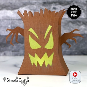 May include: A brown papercraft tree with a spooky face cut out, featuring glowing yellow eyes and a jagged mouth. The tree has outstretched arms and the text "SVG Cut File" and "Video Tutorial".