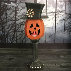 May include: A papercraft jack-o'-lantern with a black top hat and a lit candle inside. The jack-o'-lantern is on a black pedestal with white dots.