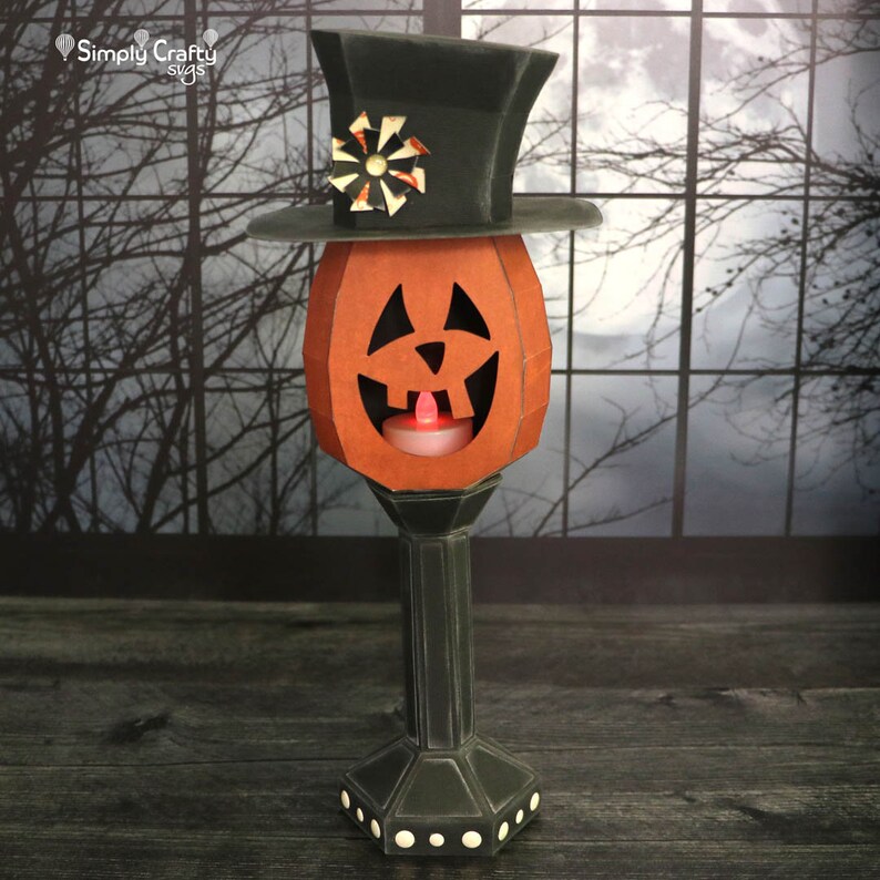 May include: A papercraft jack-o'-lantern with a black top hat and a lit candle inside. The jack-o'-lantern is on a black pedestal with white dots.