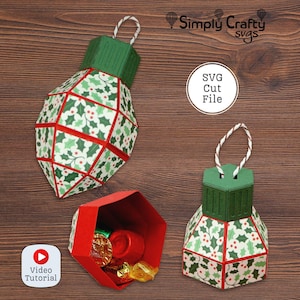 May include: Three Christmas ornament-shaped paper boxes. The boxes are green, red, and white with a holly leaf pattern. One box is open, revealing wrapped candies. The image includes the text "Simply Crafty svgs" and "SVG Cut File".
