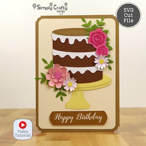 May include: A handmade birthday card featuring a chocolate cake with white frosting, adorned with pink and white paper flowers and green leaves. The card has a beige background and a brown border, with the words "Happy Birthday".