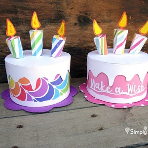 May include: Two paper birthday cakes with candles. The cake on the left has a rainbow swirl design and a purple base. The cake on the right is pink with white icing and the words "Make a Wish" on the side. Both cakes have white bases with polka dots.