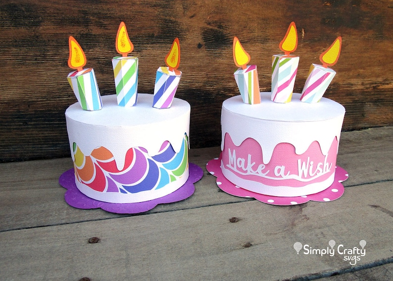 May include: Two paper birthday cakes with candles. The cake on the left has a rainbow swirl design and a purple base. The cake on the right is pink with white icing and the words "Make a Wish" on the side. Both cakes have white bases with polka dots.