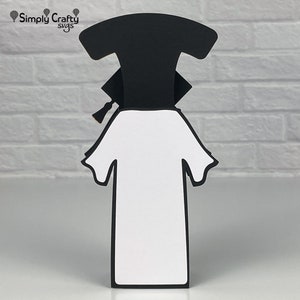 Pode incluir: Uma caixa em forma de chap&eacute;u e beca de gradua&ccedil;&atilde;o, com uma beca branca e um chap&eacute;u preto. A caixa &eacute; feita de cartolina e est&aacute; pronta para ser decorada.