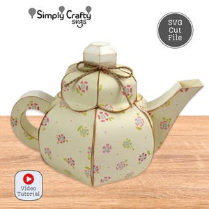 Può includere: Una teiera di carta con un motivo floreale rosa e verde su sfondo crema. La teiera ha un fiocco di spago marrone e un manico. L'immagine include il testo "Simply Crafty svgs" e "SVG Cut File".