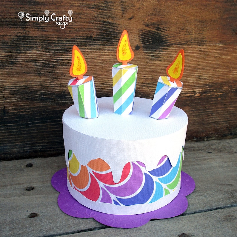 May include: A papercraft birthday cake with three colourful striped candles and a rainbow swirl design. The cake is on a purple base.