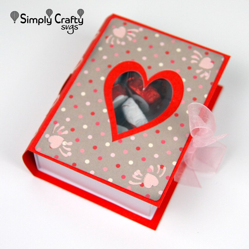 May include: A red and gray scrapbook with a heart-shaped cutout. The cover features a polka dot pattern and pink heart-shaped designs. A sheer pink ribbon is tied on the side. The text "Simply Crafty svgs" is in the upper left corner.