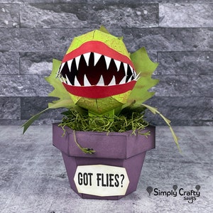 May include: A green and red papercraft model of a carnivorous plant with a toothy mouth, in a purple pot with a sign that reads "Got Flies?"