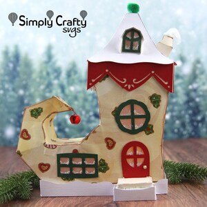 May include: A whimsical Christmas elf shoe house with a red door, green windows, and a white roof with a green pom-pom on top. The shoe is decorated with hearts and gingerbread details. "Simply Crafty svgs" is printed on the top of the image.