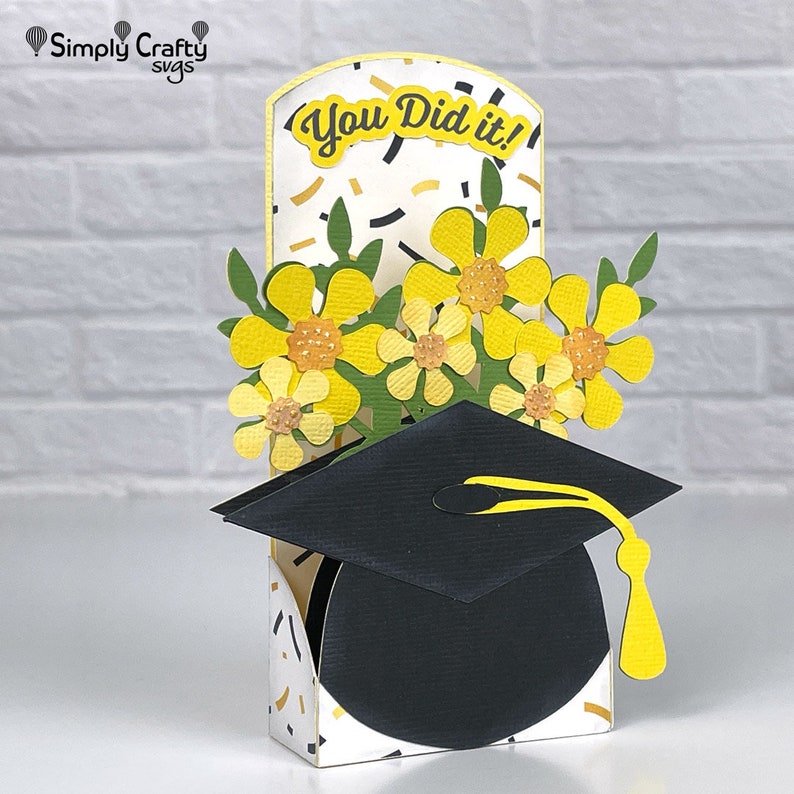 Op de afbeelding: Een afstudeerkaart met een pop-up-hoed en gele bloemen. De kaart heeft een witte achtergrond met zwarte en gouden confetti en de tekst "You Did It!"