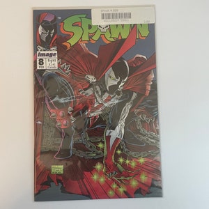Puede incluir: Una portada de cómic con un personaje de superhéroe rojo y negro con cara de calavera, de pie sobre una valla de cadena con estrellas verdes brillantes. El cómic se titula "Spawn" y es el número 8.