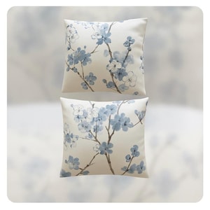 May include: Two square throw pillows with a floral design. The pillows have a cream-colored background with a pattern of blue and white flowers and branches. The pillows are stacked on top of each other.