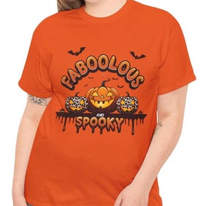 Pode incluir: Camiseta laranja com um gráfico temático de Halloween. O design apresenta as palavras "FABOOLOUS" e "SPOOKY" acima e abaixo de três abóboras. Duas abóboras têm um design de estampa de leopardo, e a abóbora central tem um rosto esculpido.