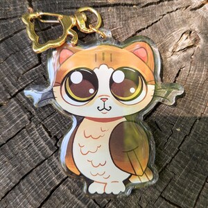May include: A clear acrylic cat-shaped keychain with a gold-colored clasp. The cat has large brown eyes, orange and brown fur, and a small pink nose. The keychain is on a wooden surface.