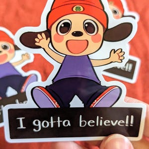 Op de afbeelding: Een sticker met een cartoonhondje met een rode muts, paars shirt en donkerblauwe broek. De sticker heeft de tekst "I gotta believe!!" op een zwarte rechthoek.