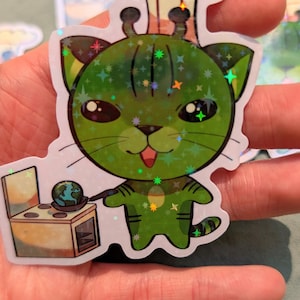Op de afbeelding: Een groene kattensticker met een koksmuts, die een spatel boven een miniatuuroven met een wereldbol erin vasthoudt. De sticker heeft een holografische, glitterende afwerking met stervormige accenten. De kat heeft grote ogen en een glimlachende uitdrukking.