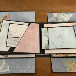 May include: A multi-layered scrapbook album featuring various paper panels in shades of light blue, soft pink, and cream. The design includes geometric pockets, fold-out flaps, and decorative collage elements with printed script and patterns.