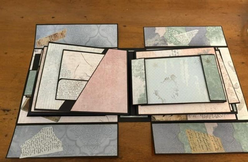 May include: A multi-layered scrapbook album featuring various paper panels in shades of light blue, soft pink, and cream. The design includes geometric pockets, fold-out flaps, and decorative collage elements with printed script and patterns.