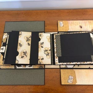 Pu&ograve; includere: Un album per ritagli artigianale a tema api e alveari, con pagine in cartoncino nero, carta fantasia color crema con illustrazioni di api ed etichette decorative. L'album aperto mostra diversi pannelli pieghevoli e tasche.