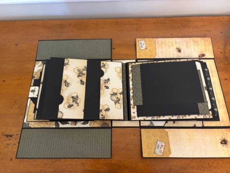 Pu&ograve; includere: Un album per ritagli artigianale a tema api e alveari, con pagine in cartoncino nero, carta fantasia color crema con illustrazioni di api ed etichette decorative. L'album aperto mostra diversi pannelli pieghevoli e tasche.