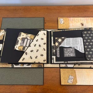Pu&ograve; includere: Un album di ritagli artigianale con pagine nere, inserti di carta con motivi di api, disegni a nido d'ape ed elementi decorativi vintage. L'album aperto mostra varie tasche, texture di carta stratificate in tonalit&agrave; nero, crema e verde oliva.